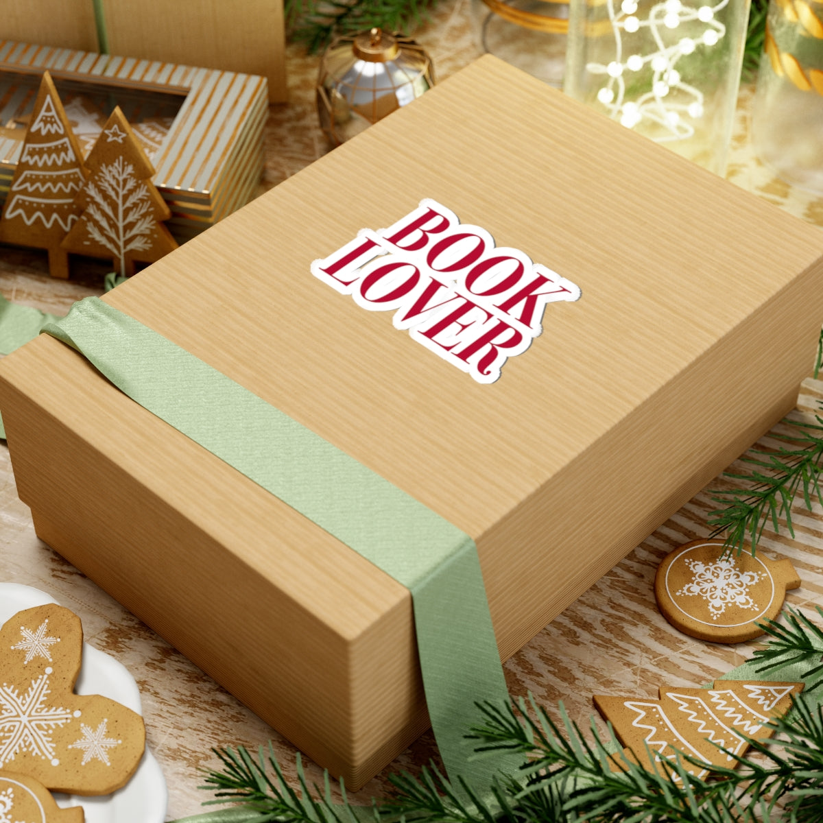 A kraft gift box with a pale green ribbon and a white sticker reading "BOOK LOVER" in bold red serif font, surrounded by holiday cookies and festive decor.
