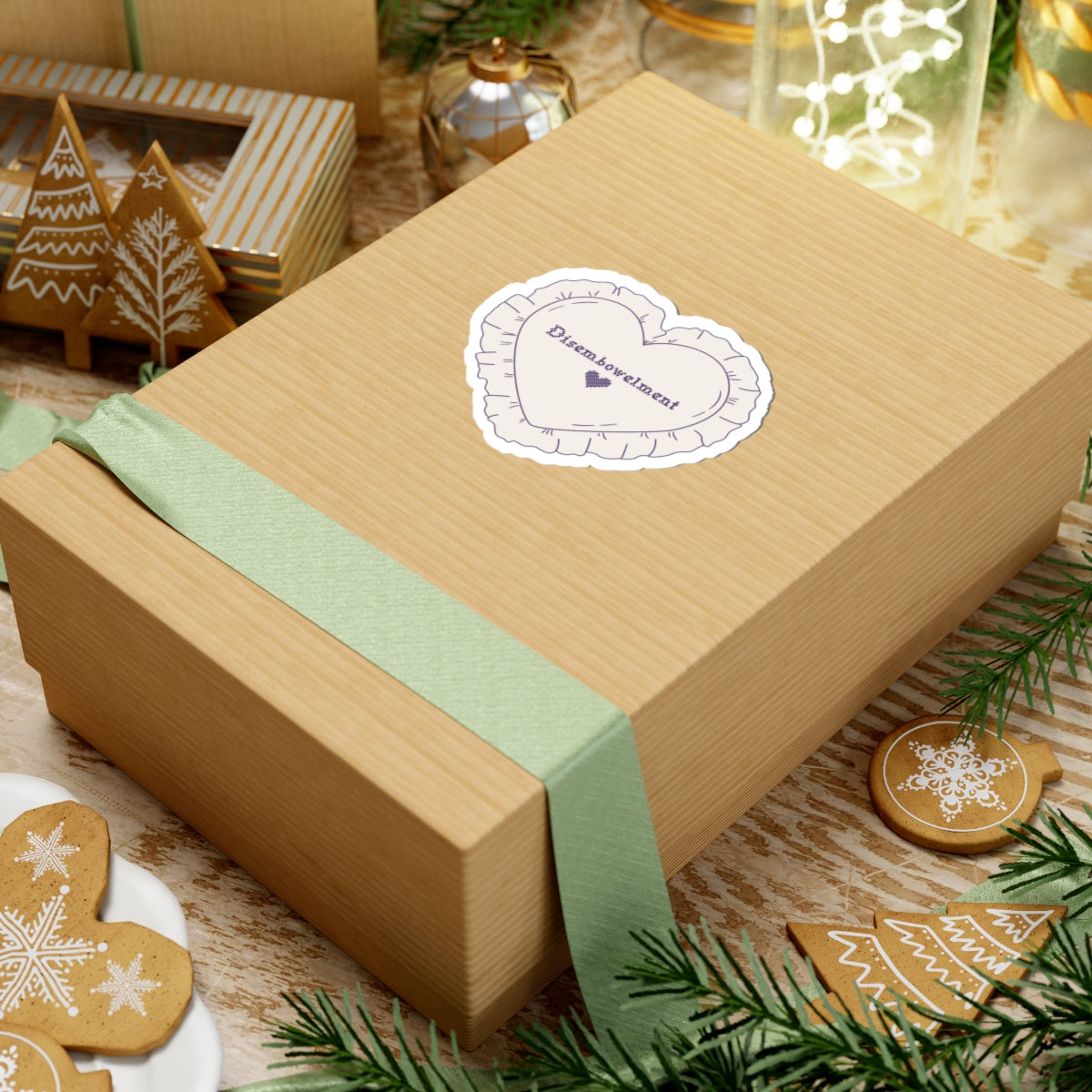 A close-up of a kraft gift box tied with a soft green ribbon featuring a thtgrlreads Disembowelment embroidery sticker from The Poison Daughter collection, surrounded by festive gingerbread cookies and holiday decor.