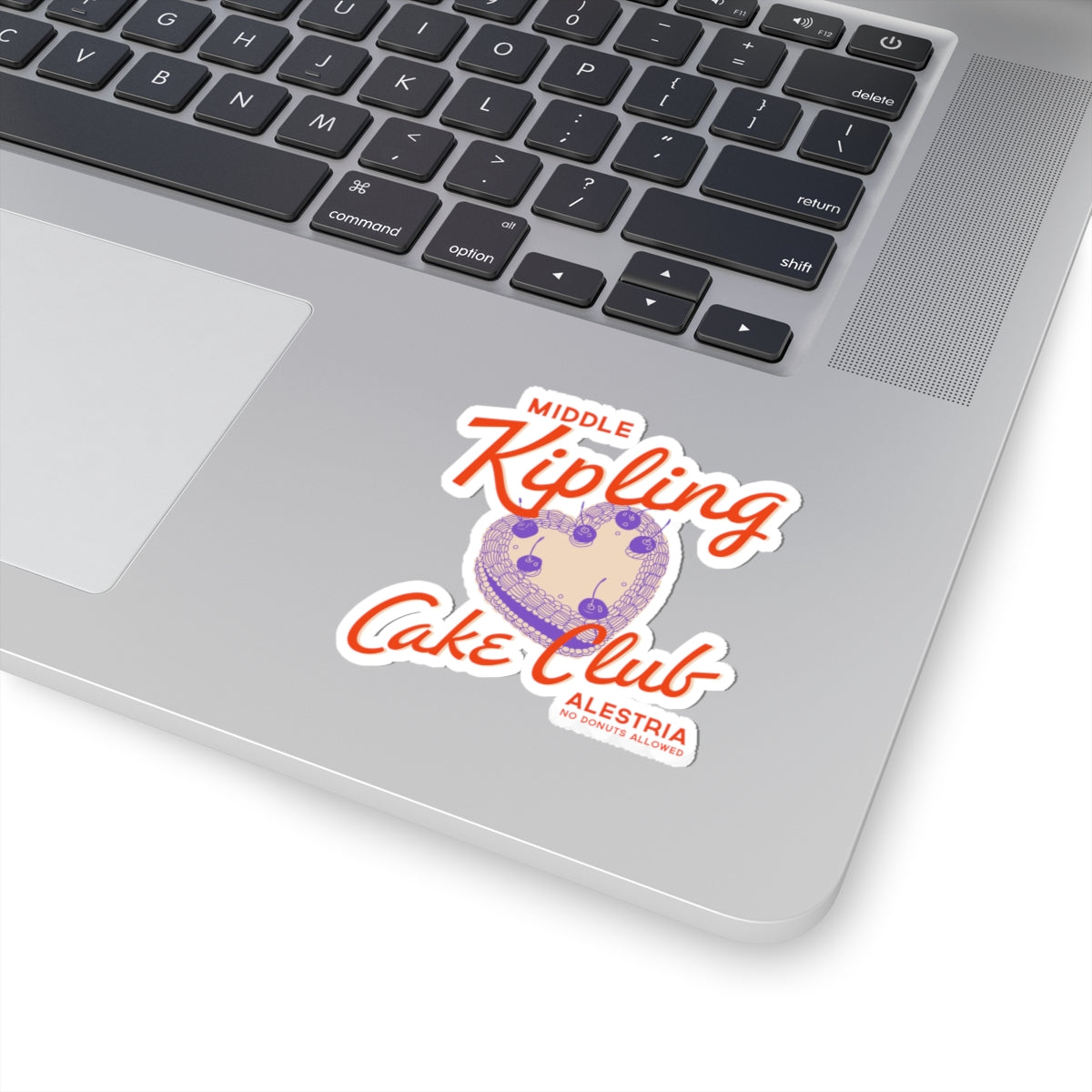 Sticker from thtgrlreads' Ruthless Boys of the Zodiac collection featuring a whimsical design with orange script text "Middle Kipling Cake Club" above a purple cake illustration, placed on a silver laptop.
