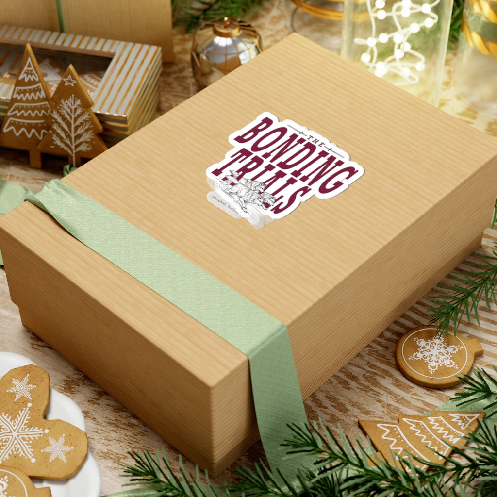 A Bonding Trials sticker from thtgrlreads featuring fantasy romance design, placed on a textured gift box with a green ribbon, surrounded by holiday decor from the Wolves of Ruin collection.