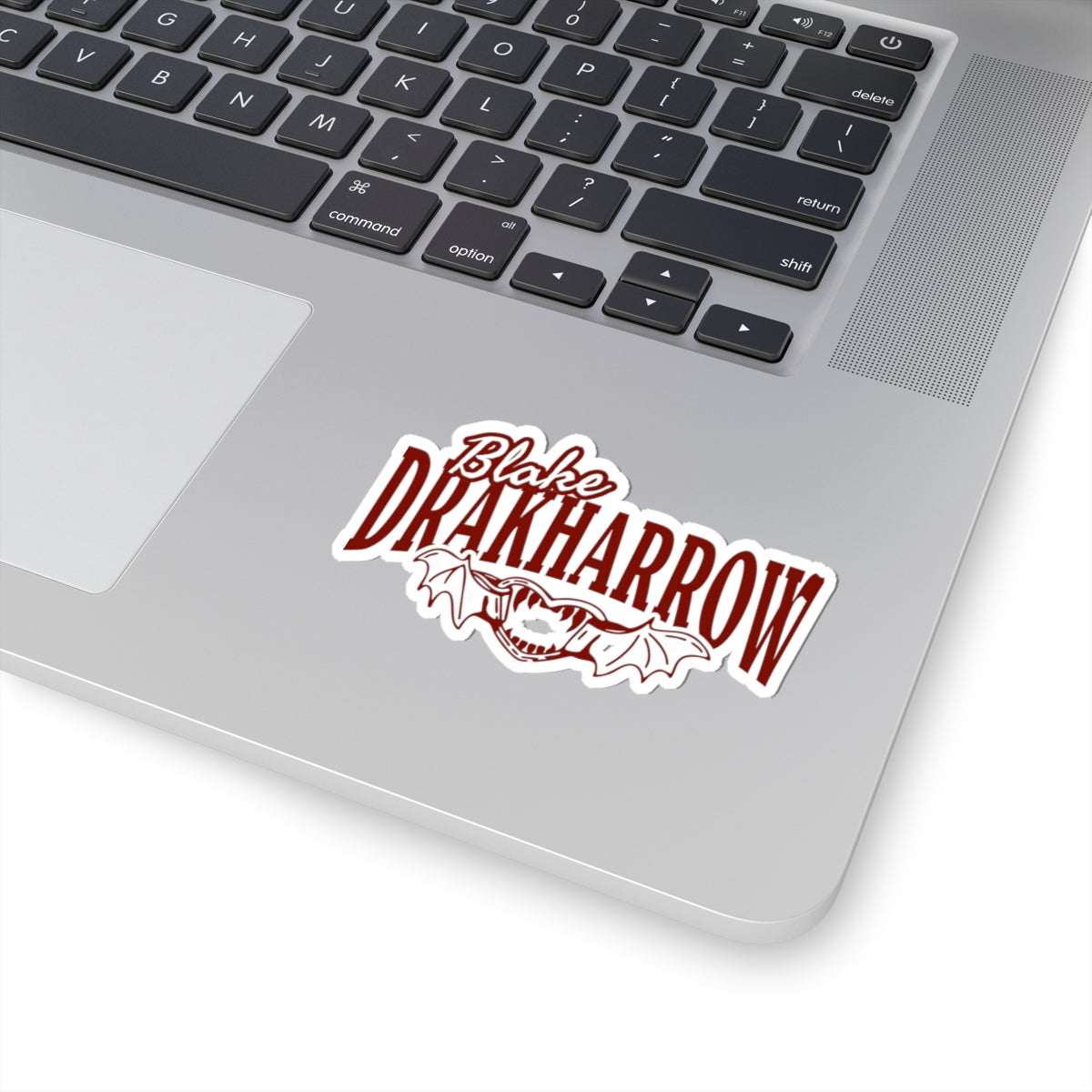 Sticker from the Bloodwing Academy collection by thtgrlreads featuring bold red gothic script text "Blake Drakharrow" with a stylized dragon wing design, shown affixed to a laptop.