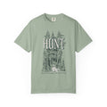 Thtgrlreads The Poison Daughter collection features a sage green Comfort Colors graphic tee with vintage wash and a detailed forest scene surrounding the large "HUNT" text in a classic font.