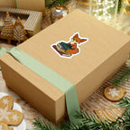 A cozy holiday gift box wrapped with a soft green ribbon, featuring a Bloodwing Academy-themed sticker from thtgrlreads showing a cute fox reading a stack of books, surrounded by festive gingerbread cookies and decorations.