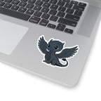 A cute black dragon chibi sticker with large eyes and outstretched wings on a silver laptop keyboard corner, featuring detailed scales and a curled tail.