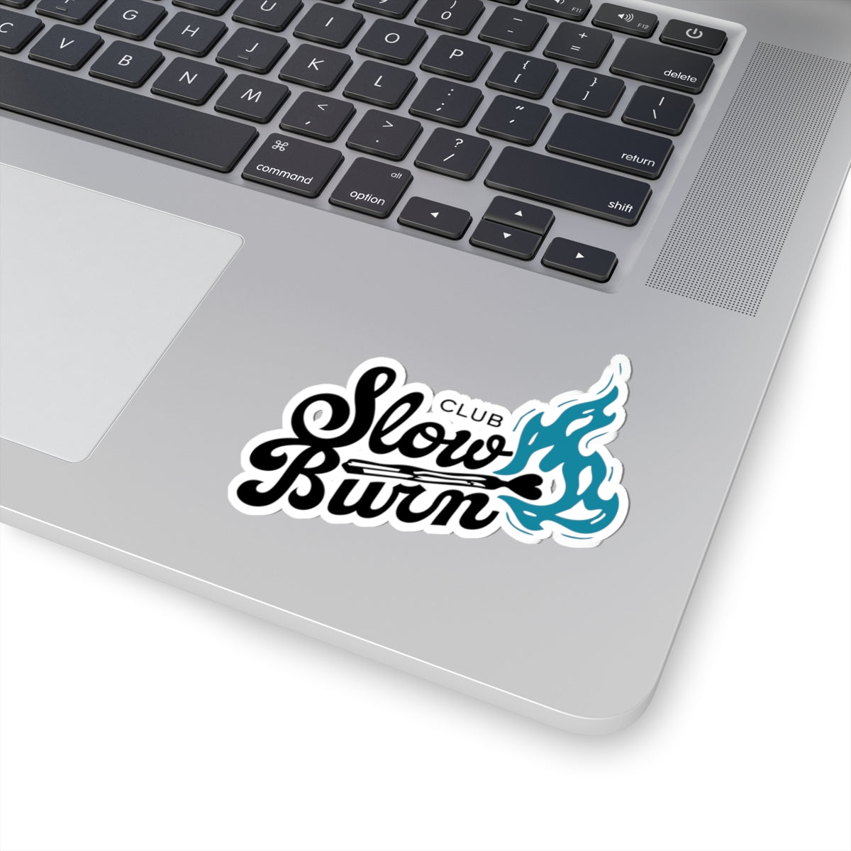 A close-up of a silver laptop keyboard area with a "Slow Burn Club" sticker featuring black cursive text and a blue flame graphic near the touchpad.
